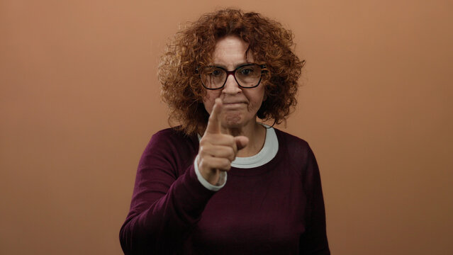 Hispanic brunette middle-aged woman performing neck-cut gesture against brown wall background with determined expression and pointing finger emphasizing seriousness.