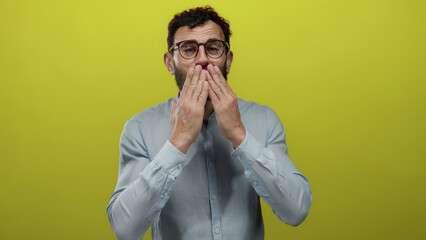 Hispanic man with beard and glasses blowing a kiss with hands towards camera against bright yellow...