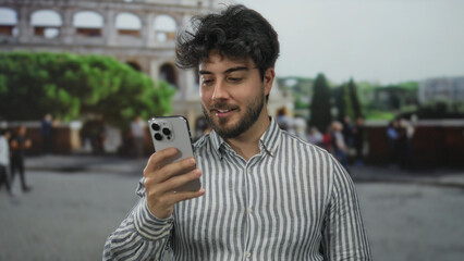 Young man with a beard using a smartphone outside the roman coliseum on a sunny day with a striped...