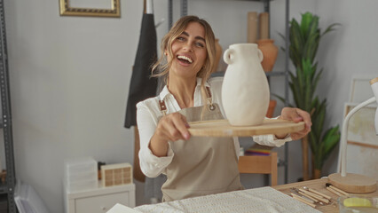 Blonde woman wearing apron holds clay vase with both hands on wooden turntable in pottery studio;...