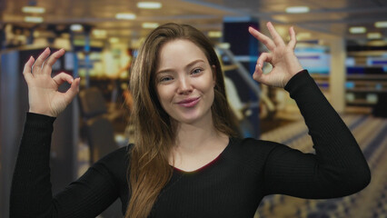 Young caucasian woman makes ok sign with both hands in a gym building surrounded by blurred workout...