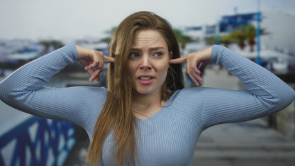 Young woman covers ears with fingers on seaside promenade while blocking out noise near a railing;...
