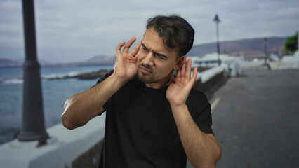Young man standing on a seaside promenade, listening intently with focus against a backdrop of sea...