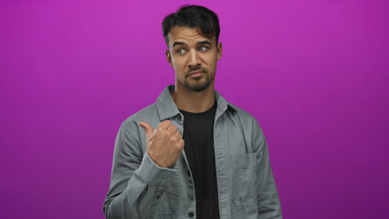 Fototapeta premium Young hispanic man in gray jacket points thumb sideways against vibrant pink background, suggesting indecision or inquiry with a confident expression.
