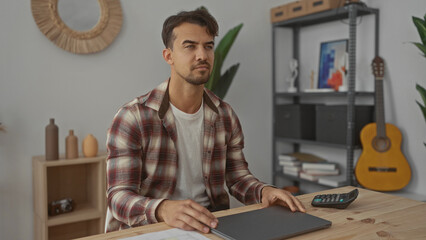 Young man working on laptop in stylish office with decor and guitar, immersed in task, embodying...