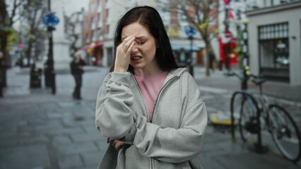 Woman in gray hoodie holding her forehead in distress on a city street with blurry buildings and bicycles in the background outdoors