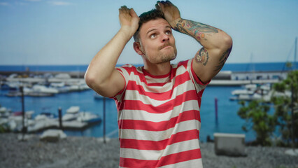 Young man in striped shirt making playful gestures at seaside port with boats and clear blue sky...