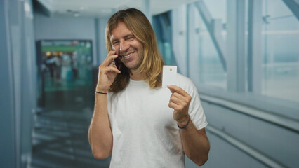 Man smiling broadly while speaking on smartphone and holding creditcard at an airport terminal;...