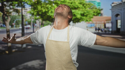 Fototapeta premium Man wearing an apron spreads out arms and tilts his head back under bright sunlight on a city street; serenity.