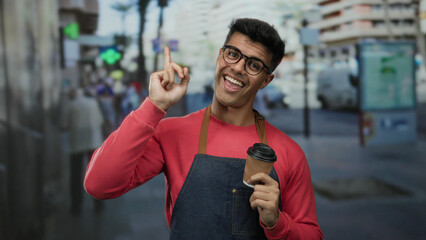 Young hispanic man wearing glasses and apron holds coffee cup on busy city street, pointing upwards...