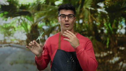 Young hispanic man in a park wearing glasses and apron holds scissors confidently, embodying a...