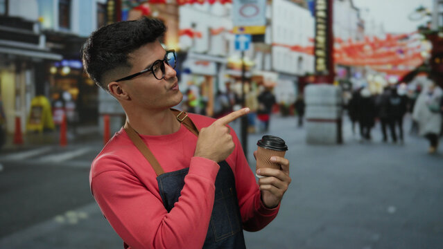 Young man with glasses holding coffee cup on bustling city street surrounded by vibrant urban setting and diverse crowd.