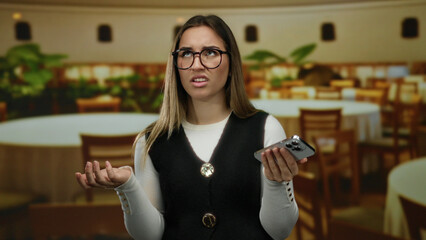 Young woman with glasses looks at her phone perplexed, standing in a cozy indoor restaurant with wooden chairs and round tables, conveying confusion and curiosity.