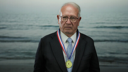 Elderly man in a business suit with a medal stands solemnly at the seaside, with the ocean waves gently rolling onto the beach in the background.