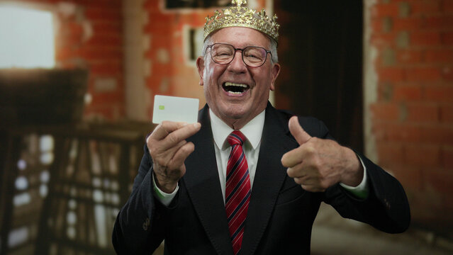 Elderly man in suit and crown gives thumbs up, holding card at indoor construction site, embodying business and triumph.