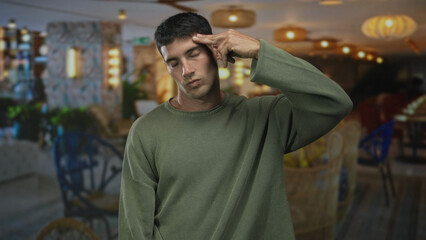 Young hispanic man in green sweater makes finger gun to head inside brightly lit building; despair...