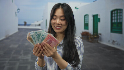 Young woman holding chinese yuan banknotes outdoors while smiling in a bright urban setting with a striped shirt and dark hair.