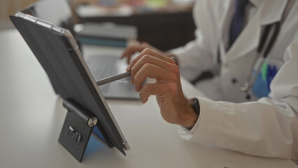 Young hispanic doctor man in white coat taps tablet screen with stylus at clinic desk beside laptop and stethoscope; focus efficiency.