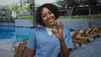 Young african american woman hand raised showing ok sign at building poolside wearing blue scrubs...