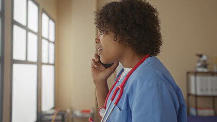 Woman doctor holds phone to ear with stethoscope around neck and gestures in clinic room; urgent concern.