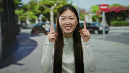 Woman smiling outdoors in urban city street setting with trees, wearing casual white attire and gesturing upwards joyfully during daytime.