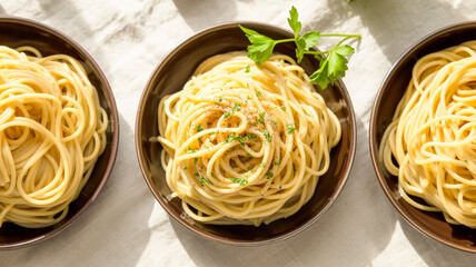 A top-down view of three identical dark brown ceramic bowls containing spaghetti pasta, arranged in a horizontal line against a light beige background