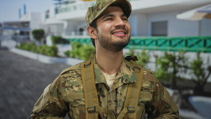 Fototapeta premium Young hispanic man in camouflage uniform shows his smiling face as he looks up on a sunlit street; pride.