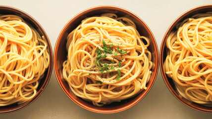 A top-down view of three identical dark brown ceramic bowls containing spaghetti pasta, arranged in a horizontal line against a light beige background