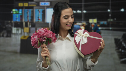 Woman smiling at airport holding a pink flower bouquet and a heart-shaped gift box, embodying...