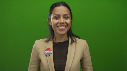 Hispanic woman smiles with 'i voted' badge on beige blazer against green background, symbolizing voting pride in united states.