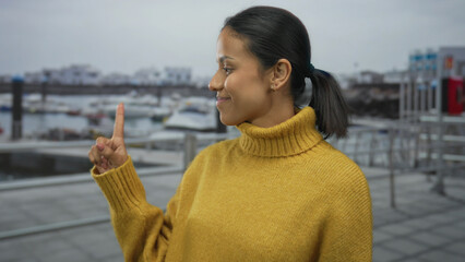 Woman in yellow sweater smiles pointing at distant boats docked at port with cloudy seaside...
