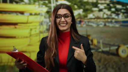 Young hispanic woman pointing to herself at a beach seaside location, holding a red clipboard...