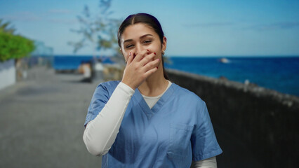 Young hispanic woman in blue scrubs laughs on a seaside promenade with ocean backdrop depicting...