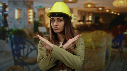 Woman architect in yellow hardhat making x gesture indoors at stylish hotel lounge expressing...