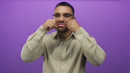 Young hispanic man wearing beige shirt rubs his eyes in mock crying gesture in purple studio set;...