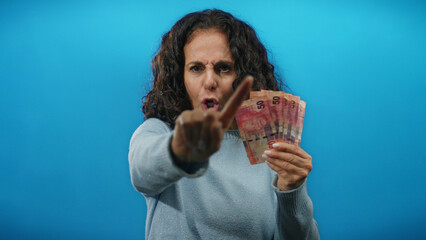 Woman gesturing angrily with finger holding south african banknotes against blue wall showing...