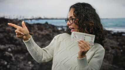 Woman happily pointing while holding uae dirham banknote on a beach with water and rocks,...