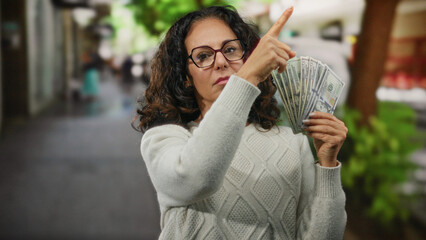 Woman holding american dollars on city street points at direction with serious expression outdoors