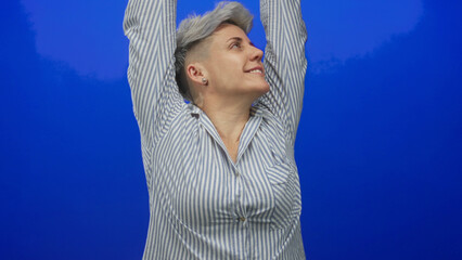 Woman in striped shirt yawning and stretching arms behind head against vivid blue studio wall;...