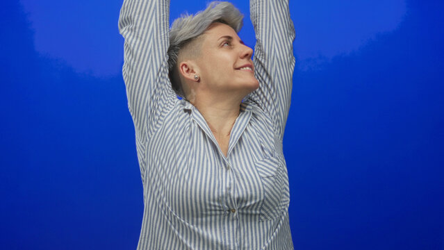 Woman in striped shirt yawning and stretching arms behind head against vivid blue studio wall; fatigue.