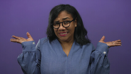 African american woman in casual blue shirt shrugging shoulders in purple studio with palms up...