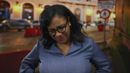 Woman wearing glasses and blue blouse stands with hands on hips on city street at night with...