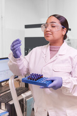young Latina college student in a white lab coat examines a biochemical sample tube with attention...