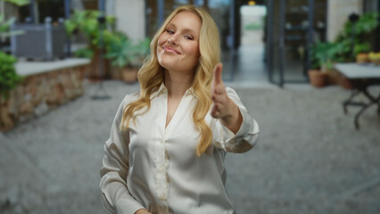 Fototapeta premium Woman smiling with extended handshake in city street background, blonde young female in white shirt outdoors surrounded by urban greenery and friendly atmosphere.