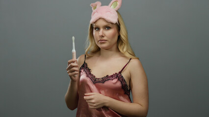 Obraz premium Young blonde woman in sleepwear holding toothbrush against grey background wearing pink sleep mask and examining toothbrush closely