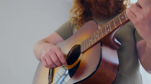 A soulful male bard with long red curls plays a heartfelt tune on acoustic guitar in his cozy room, searching for lyrics and harmony in silence.