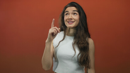 Fototapeta premium Smiling hispanic woman in white tanktop points finger up in orange studio with longhair visible and a bright grin; optimism.