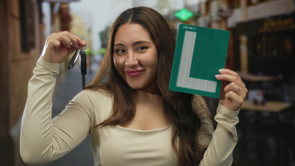Hispanic woman smiling holds dangling car keys and green learner plate outside restaurant building...