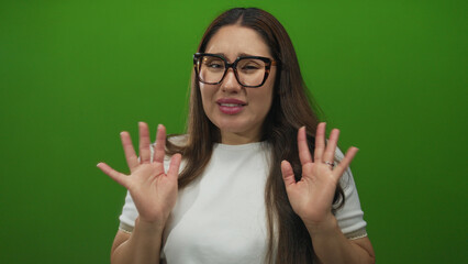 Young hispanic woman holds palms out close to chest and makes disgusted expression in green studio;...