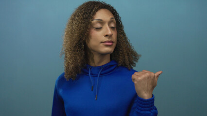 Woman pointing thumb over shoulder in blue studio with curly brown hair and hoop earrings;...